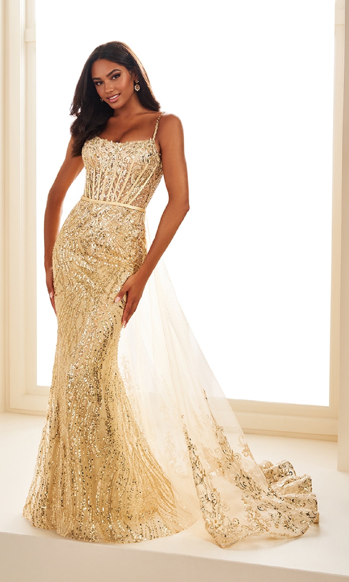 A woman poses gracefully indoors in front of a large window, wearing the Long Prom Dress: Ellie Wilde EW36075—a fitted gold sequin gown with a sheer, embroidered train.
